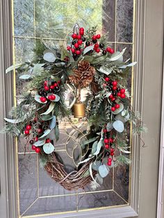 a wreath with holly, berries and pine cones hanging on a front door window sill