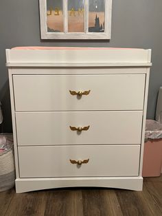 a white dresser with gold handles in a child's nursery room, next to a painting on the wall