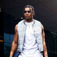 a man with dreadlocks walking down the street wearing sunglasses and a vest over his shoulders