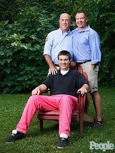 two men and a woman posing for a photo in front of some green bushes, one man is sitting on a red chair