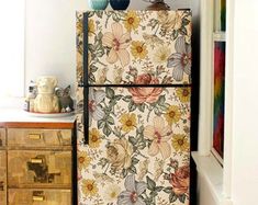 a refrigerator covered in floral wallpaper next to a dresser with drawers and vases on top