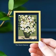 a hand holding up a small cross - stitch picture frame with flowers in the background