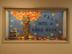 Blue paper and brown fabric are placed within a display case with bulletin board backing. Brown paper bags are crumpled to make the tree trunk, with orange and yellow leaves cut from paper. In black paper letters, the words "Fall Into a Good Book" are next to the tree. More orange and yellow paper leaves are placed all around the display to resemble falling autumn leaves. Fall Library Displays, Autumn Library, Library Magic, Fall Library, Varsity Cheerleading, Book Bulletin Board, School Library Bulletin Boards, November Bulletin Boards, School Library Decor