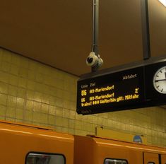 a clock hanging from the ceiling in a train station next to two yellow subway cars