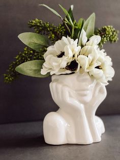 a white vase with flowers in it sitting on a table next to a gray wall