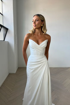 a woman in a white wedding dress standing by a window with her hands on her hips