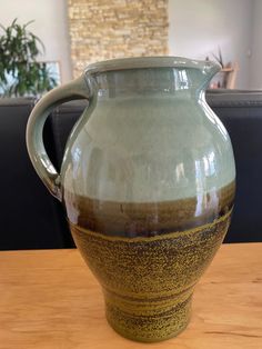 a green and brown vase sitting on top of a wooden table next to a plant