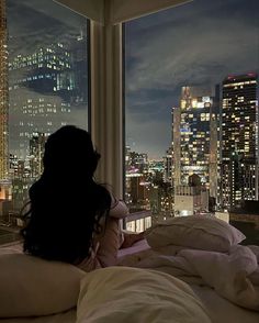 a woman is sitting in bed looking out the window at the city lights and skyscrapers