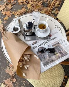 an outdoor table with newspaper, coffee cups and glasses on it in the fall leaves