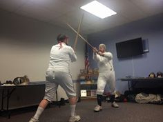 two people in white uniforms are playing with a baseball bat and another person is watching