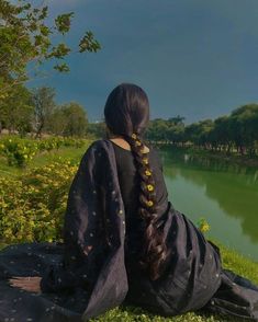 a woman with long hair sitting on the grass looking out over a body of water