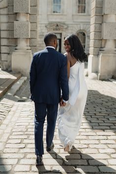 a bride and groom walking down the street in front of an old stone building holding hands