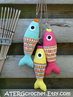 three crocheted fish hanging from hooks on a wooden fence with rakes in the background