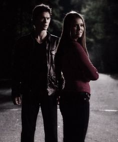 a man and woman standing next to each other on the street in the dark with trees behind them