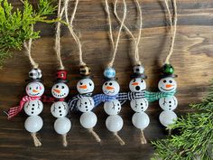 five snowman ornaments hanging from twine on a wooden table next to evergreen branches