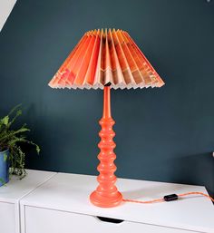 an orange table lamp sitting on top of a white dresser next to a plant and blue wall