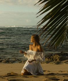 a woman sitting on top of a sandy beach next to the ocean wearing a white dress