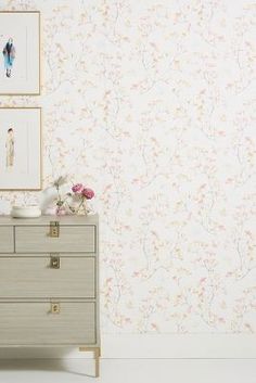 a dresser with flowers on it in front of a wallpapered background and framed pictures