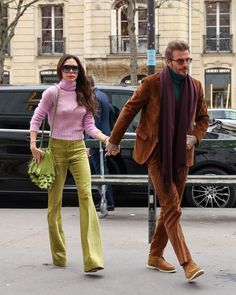 a man and woman are walking down the street holding hands while wearing green pants, pink sweater and brown boots