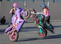 two people on motorbikes doing tricks in the street with one person wearing pink and blue