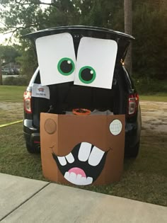 an open trunk with eyes and mouth on the grass next to a car that is parked in front of it