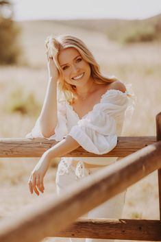 a beautiful blonde woman leaning on a wooden rail in a field wearing a white dress