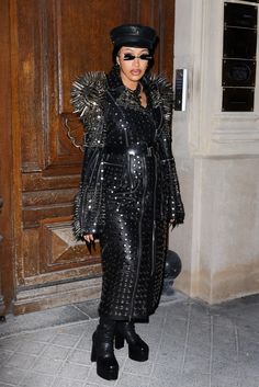 a woman in black outfit and hat standing next to a wooden door with large metal spikes on it