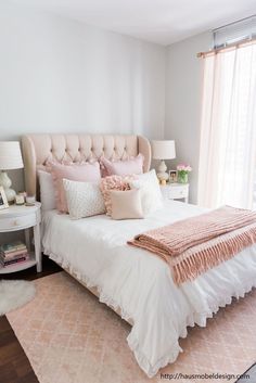 a white bed with pink pillows and blankets on it in a bedroom next to a window