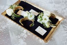 three boutonnieres with white flowers are in a box on a lace tablecloth