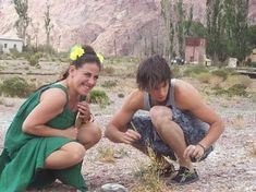 two people crouching in the desert with one holding a plant and the other eating something