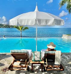 two lounge chairs under an umbrella next to a pool with the ocean in the background