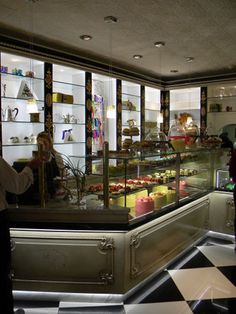 two people standing in front of a display case filled with cakes and pastries at a bakery