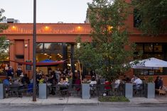 people sitting at tables in front of an orange building with trees and umbrellas on the sidewalk