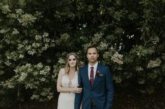 a man and woman standing next to each other in front of some bushes with white flowers