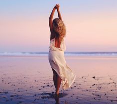 a woman in a white dress is walking on the beach with her arms raised up