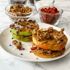 an apple sandwich on a white plate with other ingredients in bowls behind it and two glass bowls filled with granola