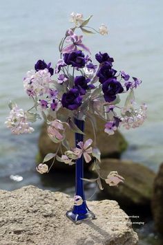 a blue vase filled with purple and white flowers on top of a rock near the ocean