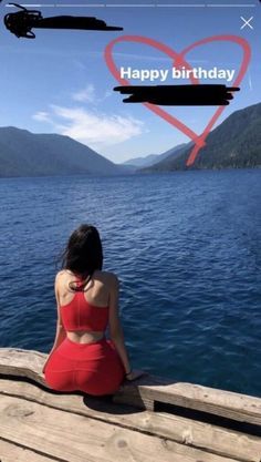 a woman in a red dress sitting on a dock looking out at the water with a helicopter flying over her head