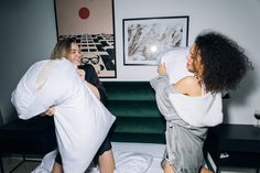 two women sitting on a bed with pillows in their hands and one is holding a pillow