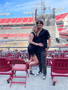 a man and woman posing for a photo in front of an empty stadium with red chairs