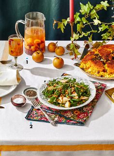 a table topped with plates of food next to a vase filled with oranges and apples