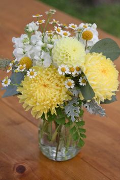 a glass vase filled with yellow and white flowers on top of a wooden table next to grass