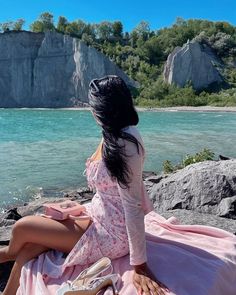 a woman in a dress sitting on the rocks by the water with her feet up