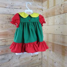 a green dress with red and yellow ruffles hanging on a wooden wall next to a hanger