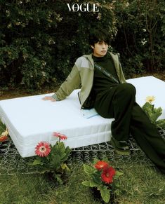 a young man sitting on top of a mattress in the middle of some grass and flowers