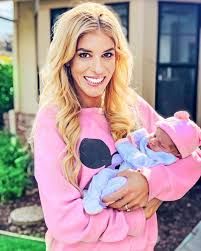 a woman holding a baby wearing a pink sweater and blue pants in front of a house