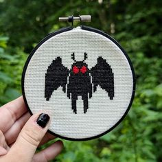 a hand holding up a cross stitched bat ornament in front of some trees