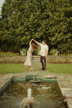 a man and woman are dancing in front of a water fountain with trees behind them