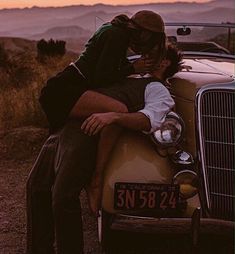 a man and woman sitting on the hood of a car in front of a sunset