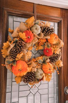 a wreath with pumpkins, pine cones and leaves hanging on the front door window